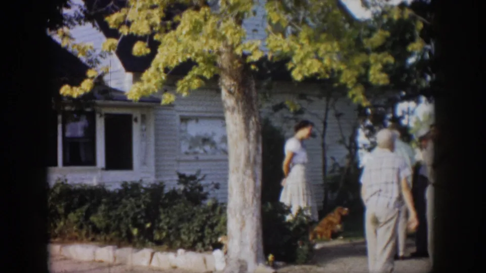 1959 Colorado home-movie frame showing a man walking toward a woman and dog in front of a white clapboard house.