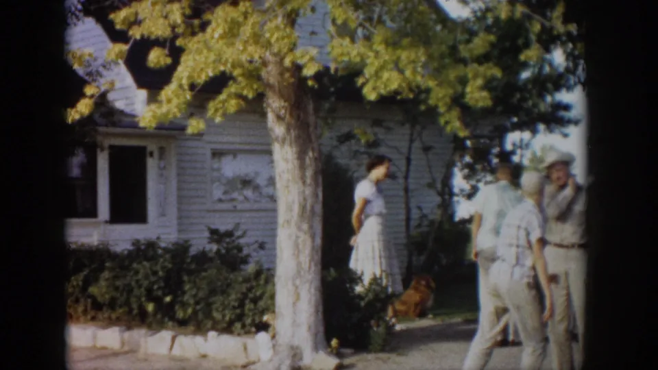 1959 Colorado home-movie frame with two men joining a woman and dog in the yard outside a white house.