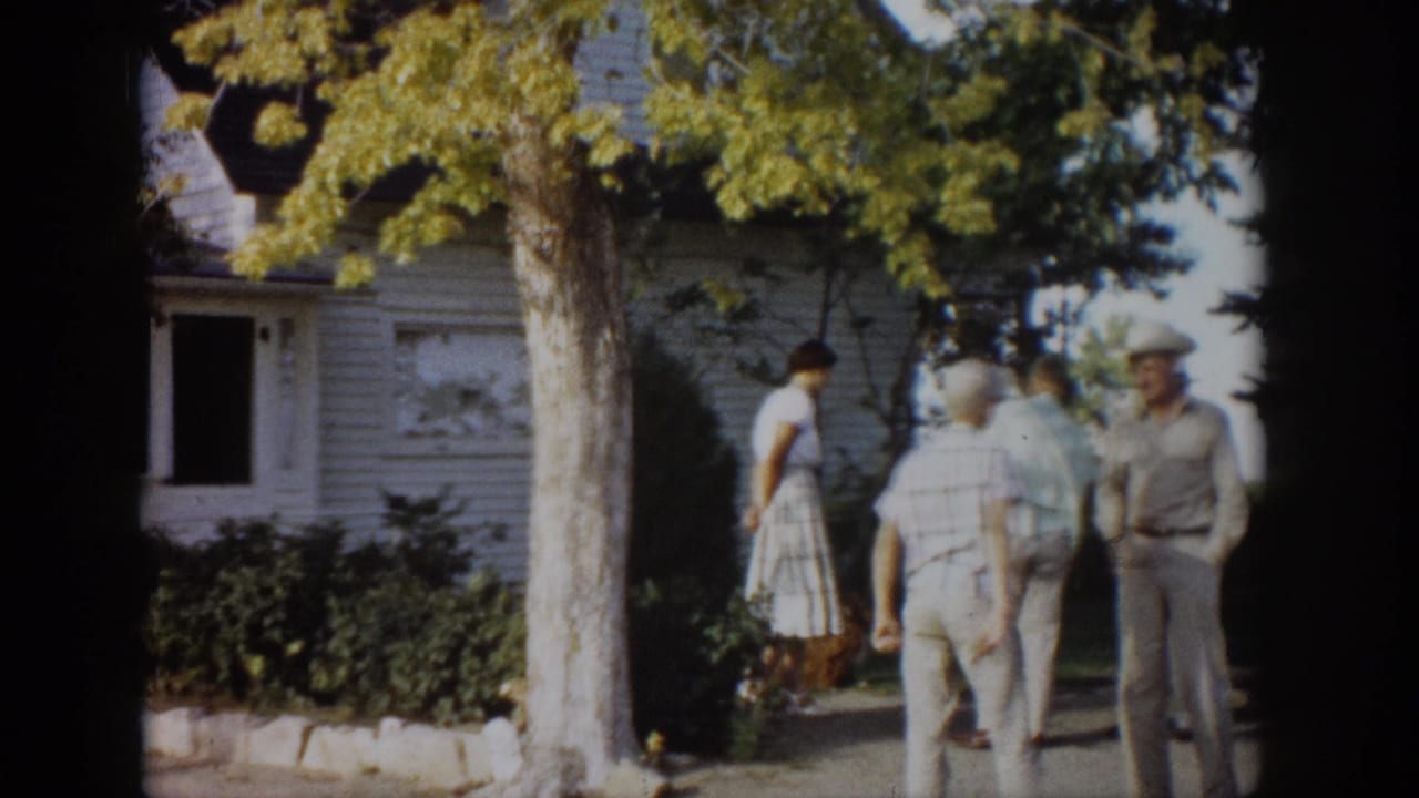 Adults stand and walk near a white house shaded by trees.