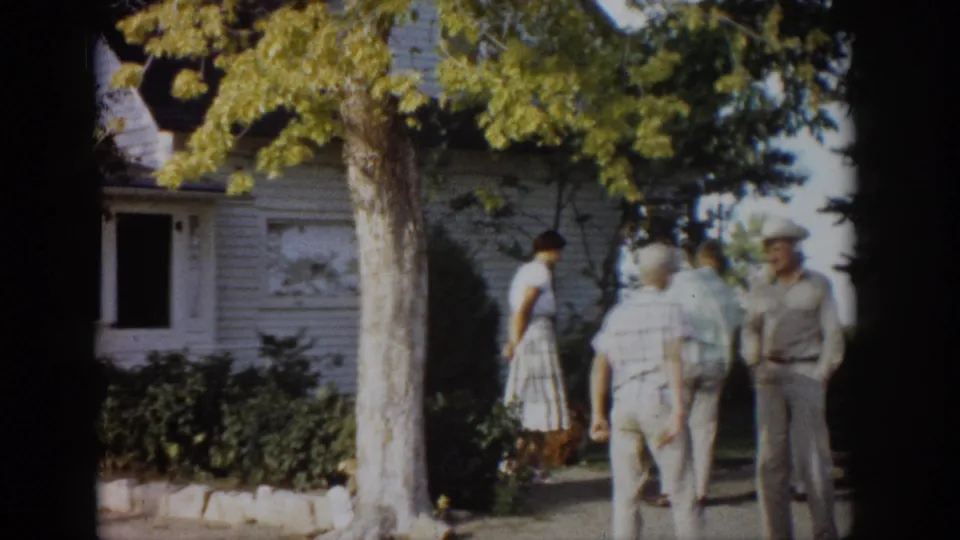 1959 Colorado home-movie frame of adults in light clothing gathered outside a white clapboard house under leafy trees.