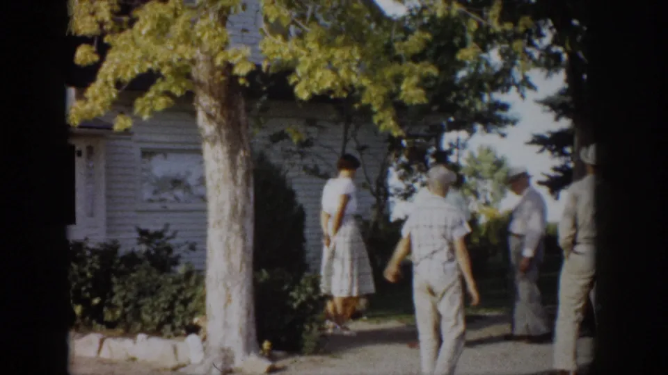 Adults in hats and light summer clothes move along a path outside a white house in the Colorado home-movie scene.