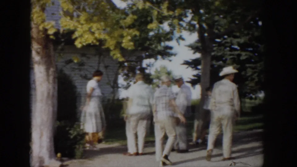 Adults in mid-century clothing walk away from the camera along a shaded path near trees and a house exterior.