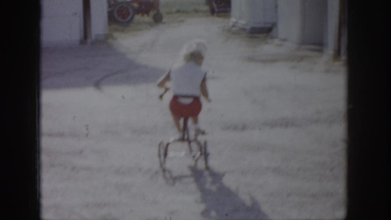 In 1959 Colorado home-movie footage, a girl in a white top and red shorts rides a bicycle away from the camera near outbuildings.