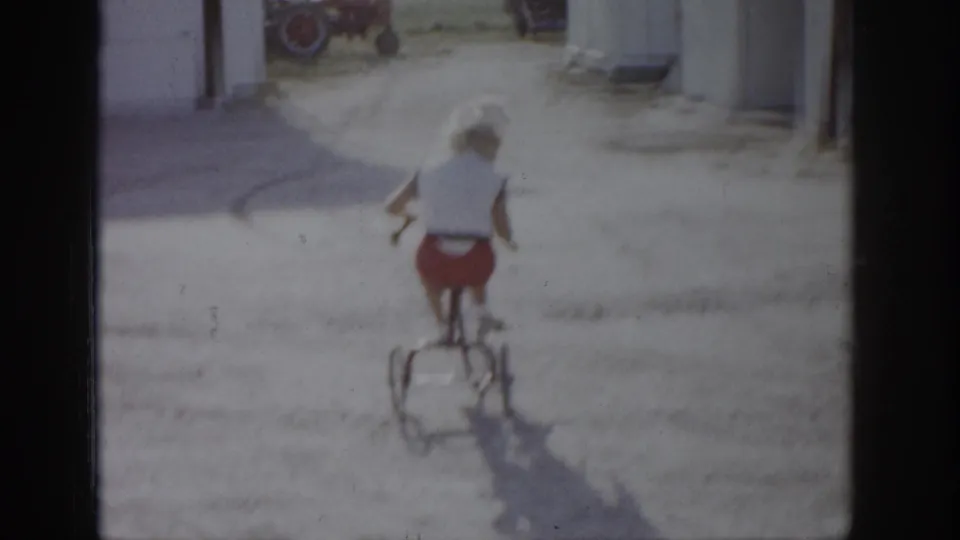 In 1959 Colorado home-movie footage, a girl in a white top and red shorts rides a bicycle away from the camera near outbuildings.