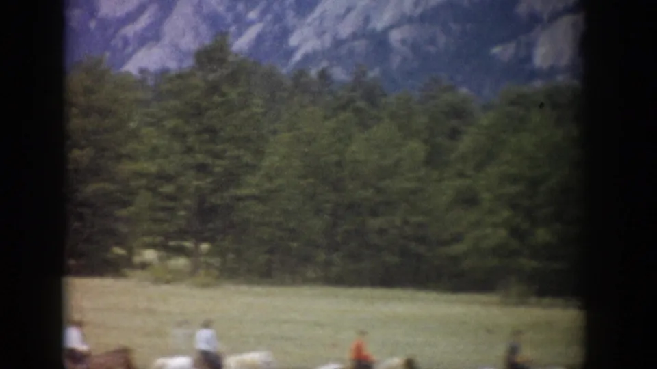 Colorado mountain and forest view with distant horseback riders crossing a grassy foreground in soft 8mm color footage.