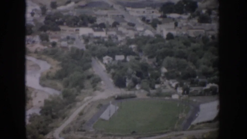 1959 Colorado aerial home-movie frame with a winding river, roads, clustered buildings, trees, and a green athletic field with a track.