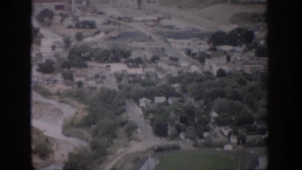 1959 Colorado aerial frame looking over roads, homes, trees, a river bend, and a green athletic field near a running track.
