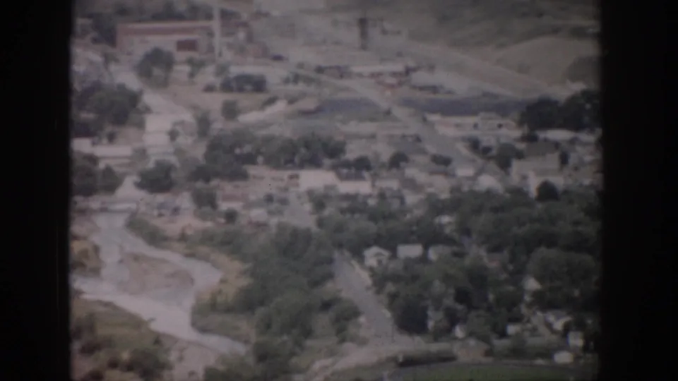 1959 Colorado 8mm aerial frame showing town buildings, a curving road, trees, a river corridor, and a green athletic field with track.
