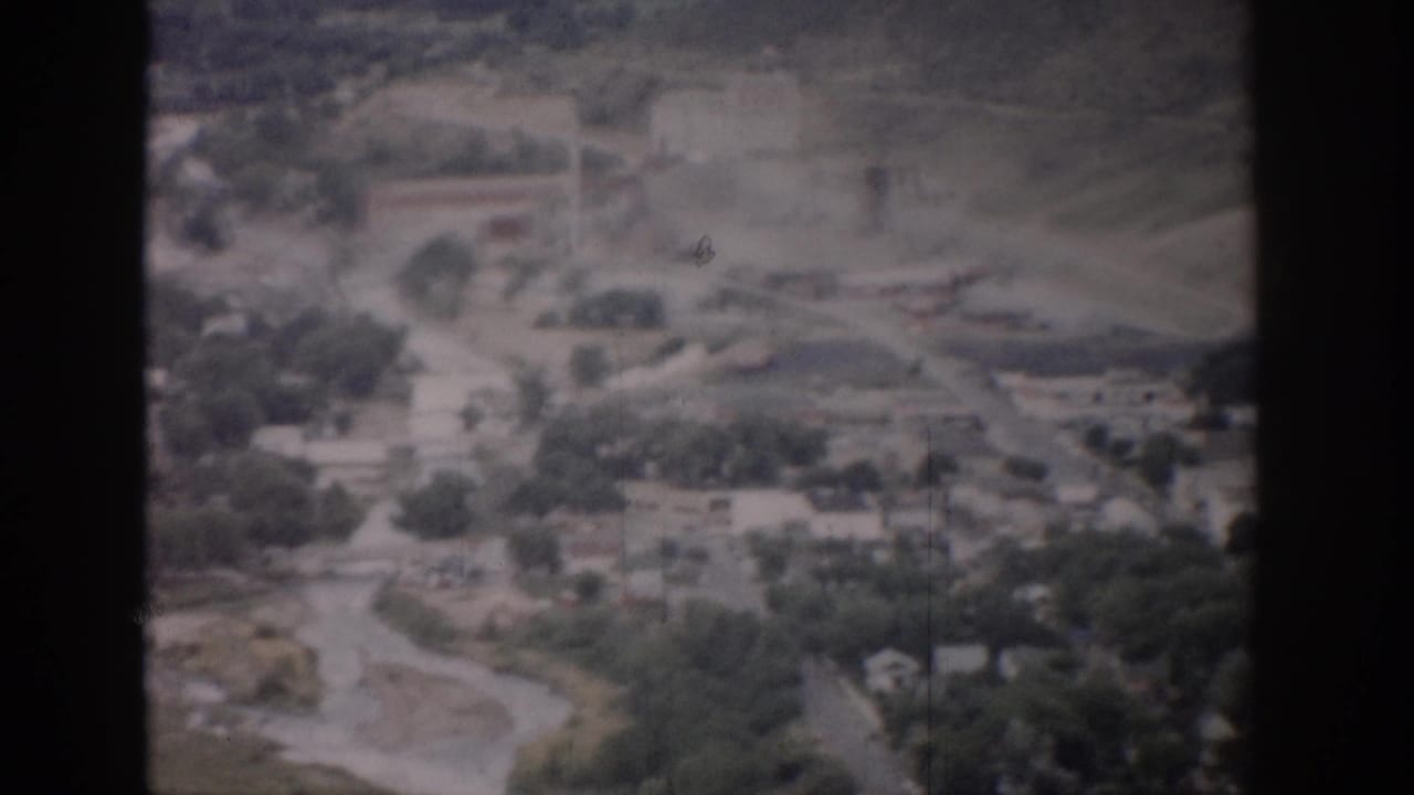1959 Colorado aerial frame with curving roads, tree cover, scattered houses, and a larger rectangular building in the distance.