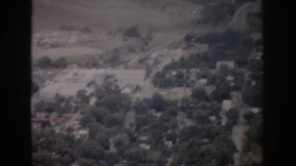 1959 Colorado 8mm frame showing a town edge from above, with green trees, roads, scattered buildings, and pale open land.
