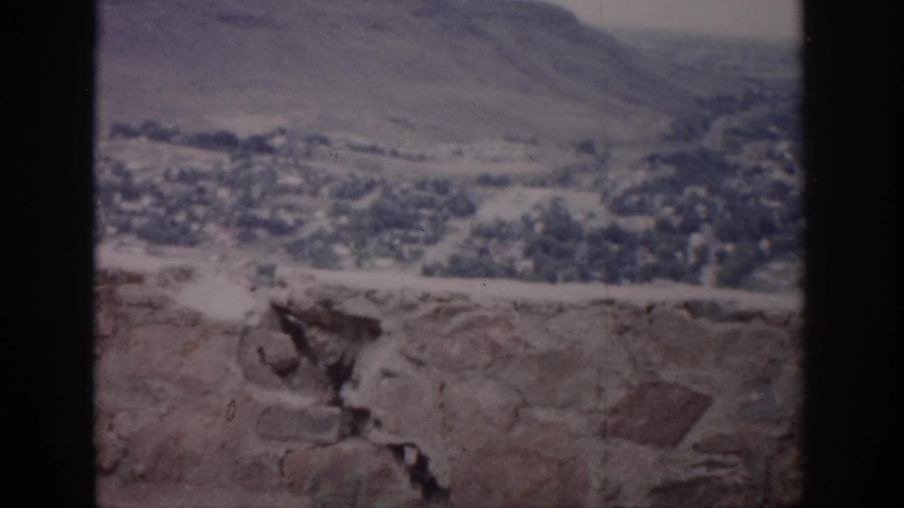 1959 Colorado 8mm frame showing a distant valley town, dry hills, hazy sky, and a rough stone ledge within dark vertical framing.
