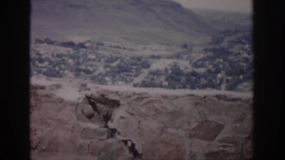1959 Colorado 8mm frame showing a distant valley town, dry hills, hazy sky, and a rough stone ledge within dark vertical framing.