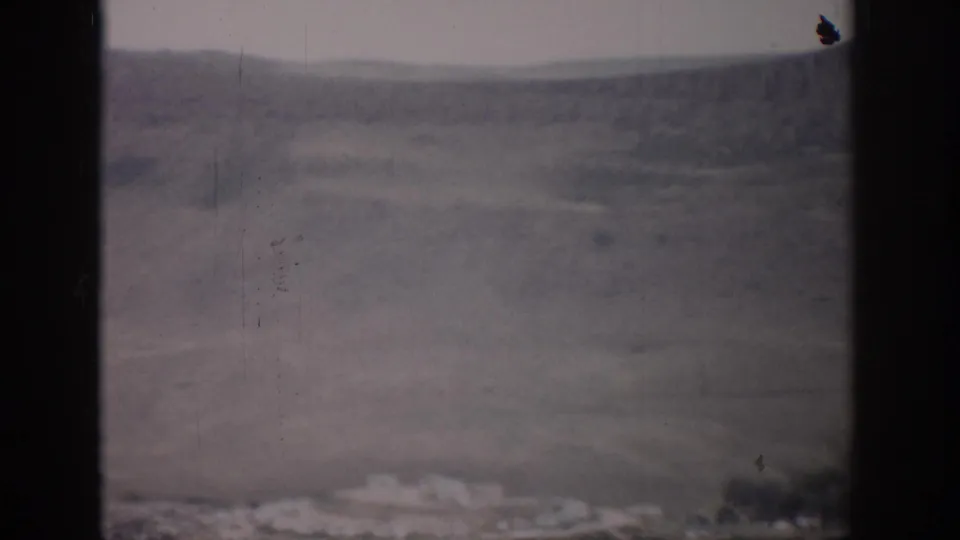 1959 Colorado 8mm frame showing a broad dry valley, layered hills, pale sky, and small distant structures near the bottom edge.