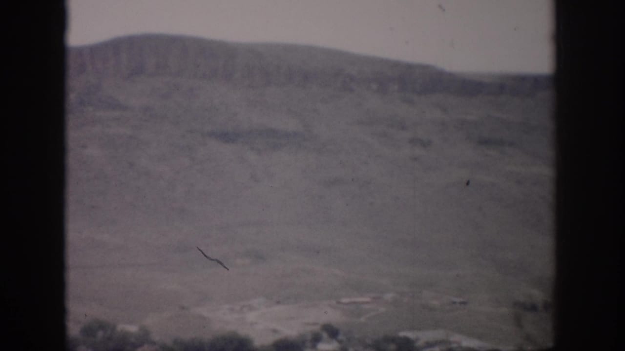 1959 Colorado 8mm frame showing a dry butte above a small valley settlement, viewed through dark vertical edges.