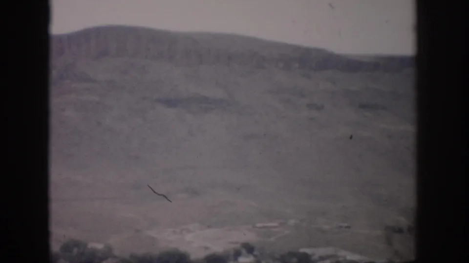 1959 Colorado 8mm frame showing a dry butte above a small valley settlement, viewed through dark vertical edges.