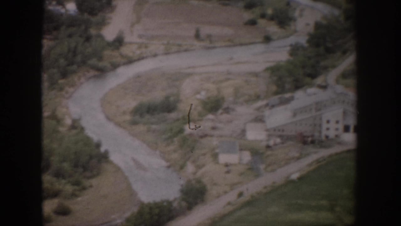 Aerial view of a winding river or road, open fields, trees, and a large building below.