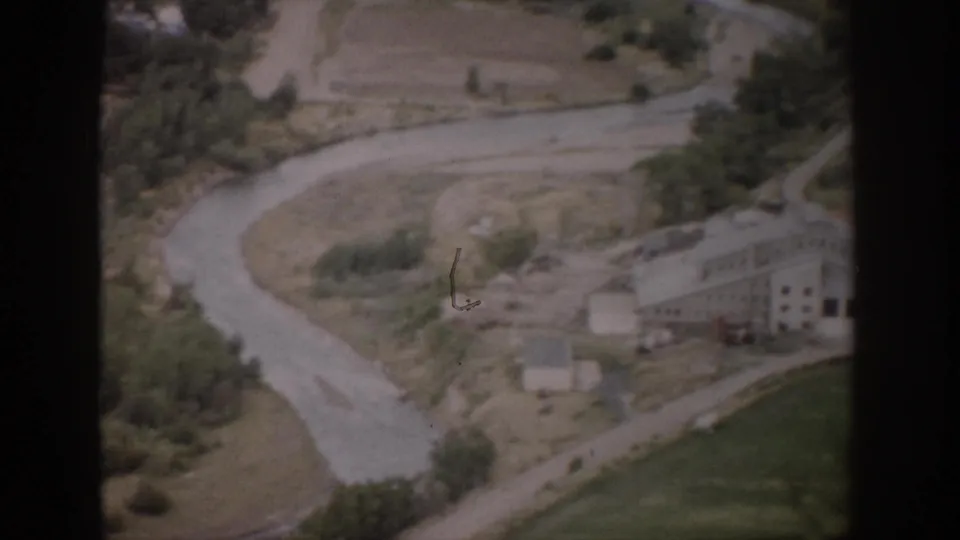 1959 Colorado 8mm aerial view over a rural valley with a curving waterway or road, dry fields, trees, and a light-colored building.