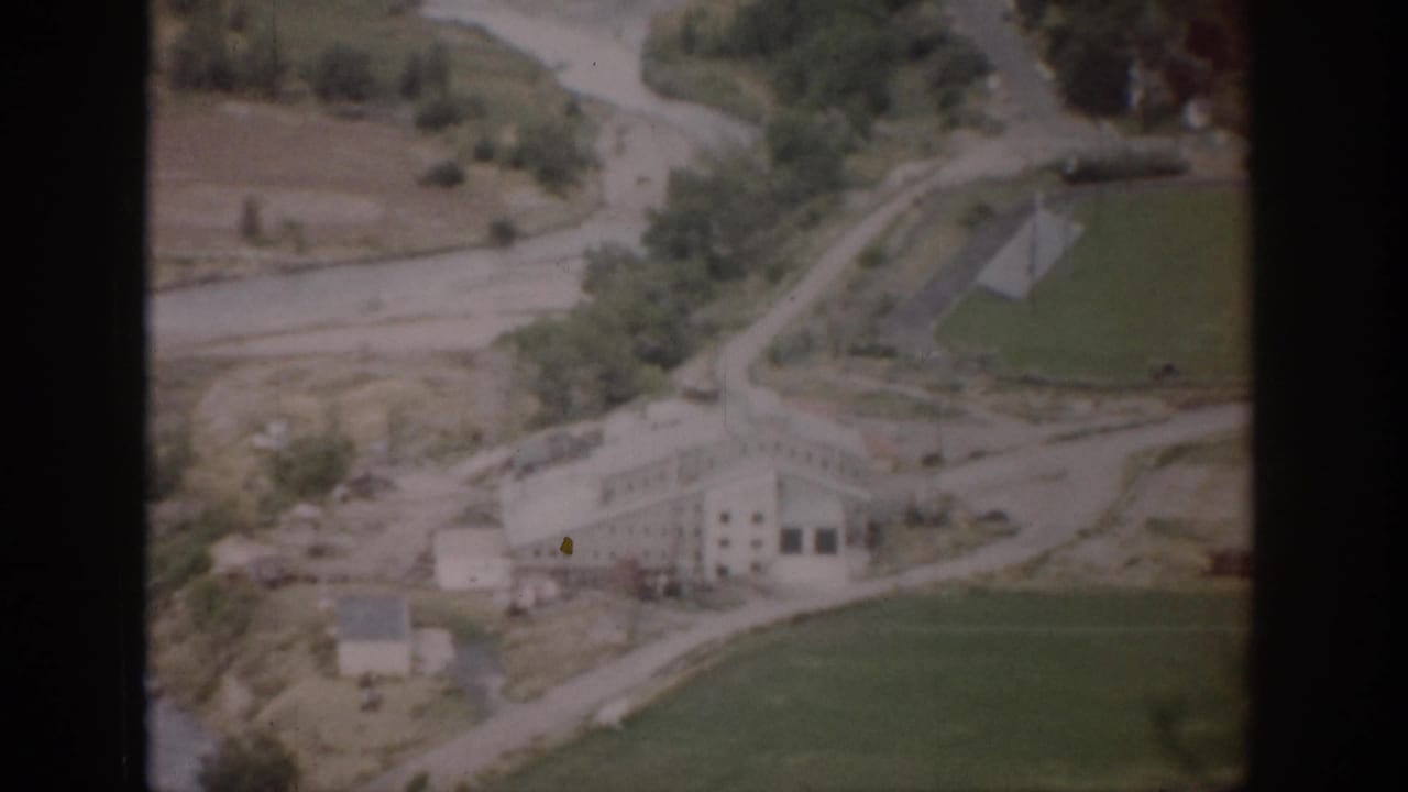 1959 Colorado aerial frame showing a light-colored building, green field, winding road or waterway, trees, and surrounding rural terrain.