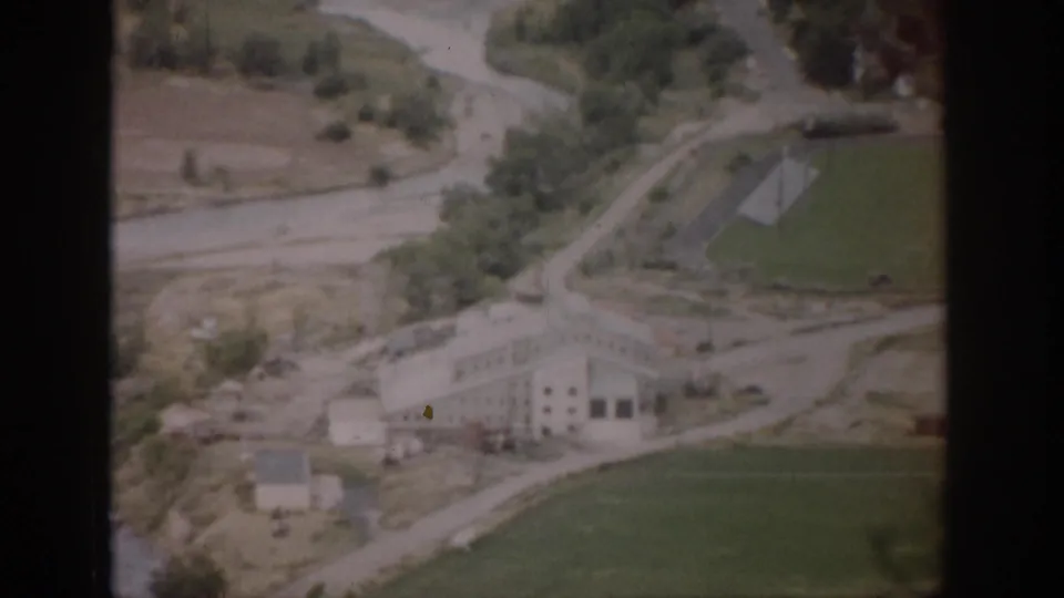 1959 Colorado aerial frame showing a light-colored building, green field, winding road or waterway, trees, and surrounding rural terrain.
