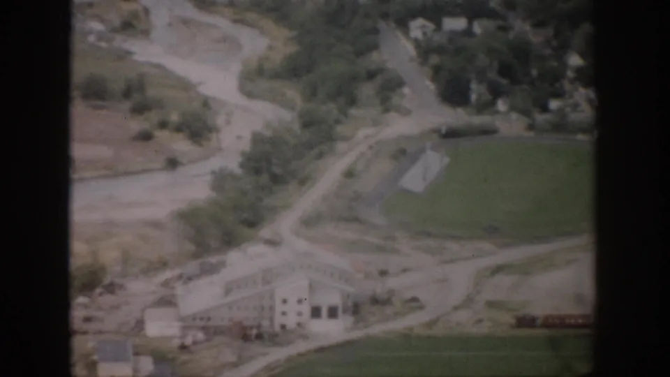 1959 Colorado 8mm aerial view with a large rural building, green field, scattered trees, and a winding route through the valley.