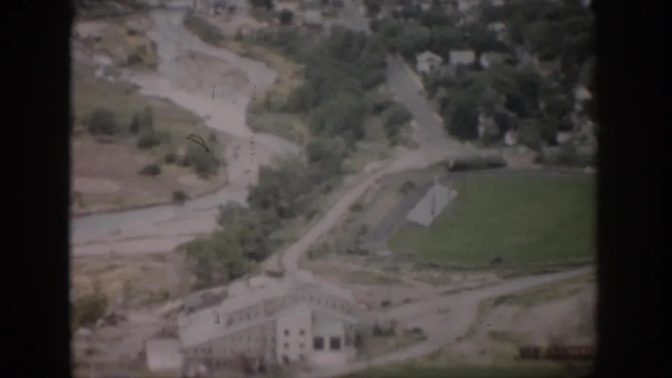 1959 Colorado aerial frame with a rural building at lower center, green field, tree-lined route, open ground, and valley roads or waterways.
