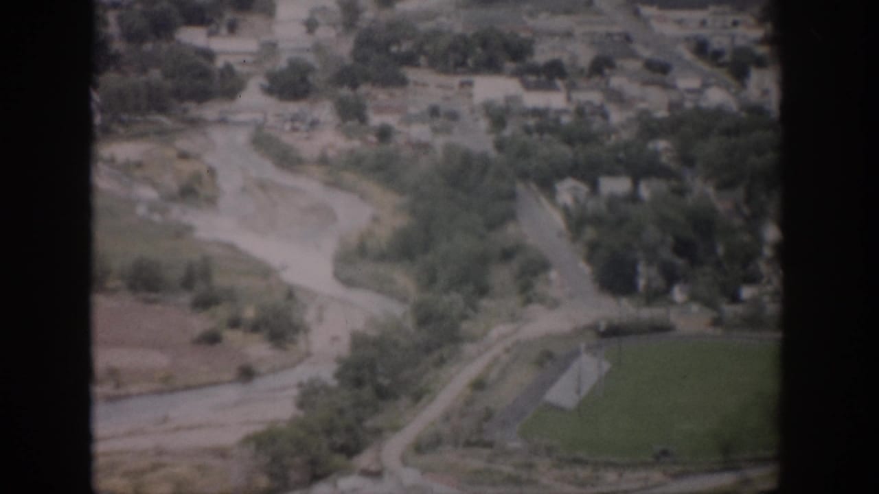 1959 Colorado aerial home-movie frame showing a river corridor, curving roads, scattered buildings, trees, and a green field.