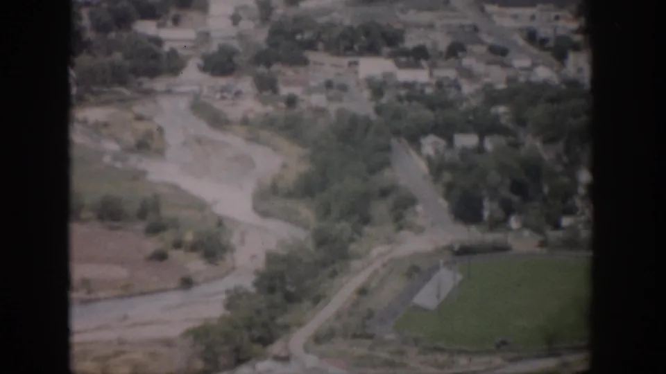 1959 Colorado aerial home-movie frame showing a river corridor, curving roads, scattered buildings, trees, and a green field.