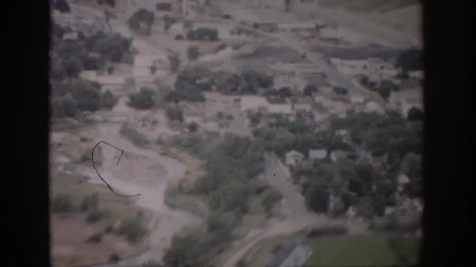 1959 Colorado aerial view with a winding watercourse, roadside trees, scattered homes, and open terrain seen through faded 8mm film.