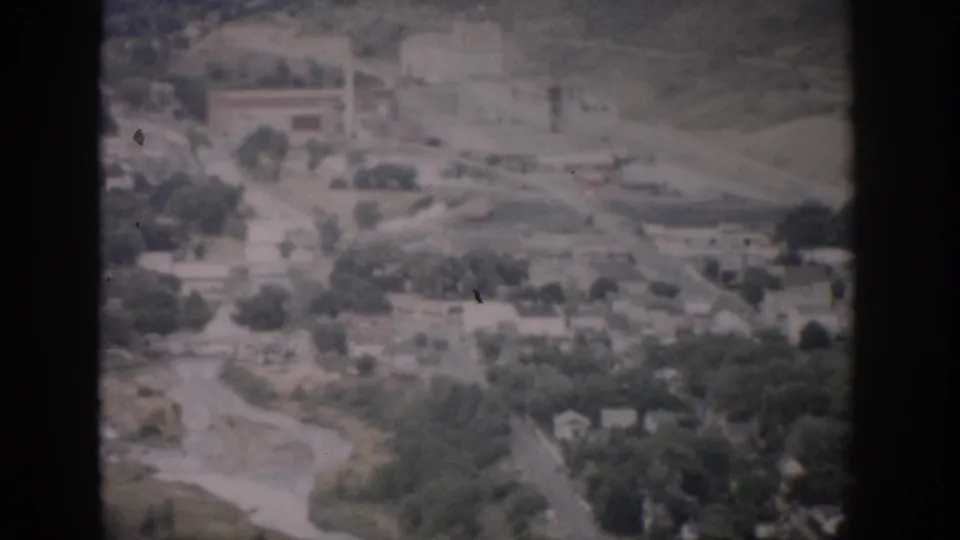 1959 Colorado aerial frame with soft focus showing roads, scattered buildings, tree cover, open ground, and a watercourse below.
