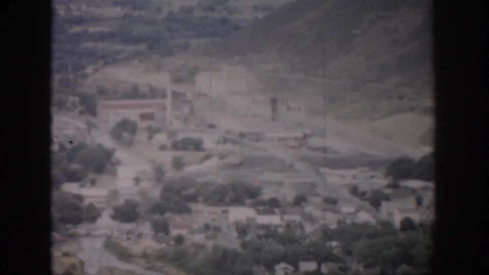 1959 Colorado home-movie aerial frame showing a hillside edge, roads, trees, scattered buildings, and open town land.