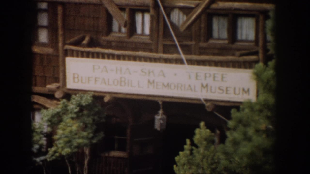 1959 Colorado 8mm frame showing the wooden Buffalo Bill Memorial Museum exterior, a large sign and green trees in soft focus.