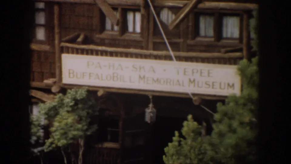 1959 Colorado 8mm frame showing the wooden Buffalo Bill Memorial Museum exterior, a large sign and green trees in soft focus.