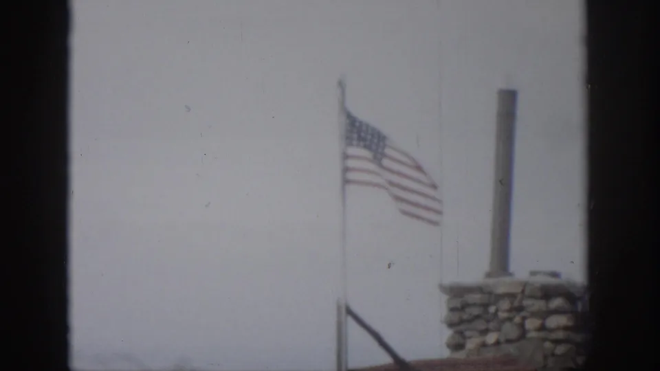 1959 Colorado 8mm frame with an American flag on a pole beside a stone chimney and vertical metal pipe under an overcast sky.