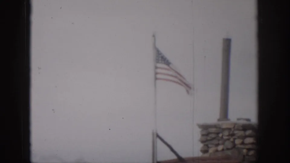 Soft 1959 Colorado home-movie frame of a weathered American flag flying beside a stone chimney and vertical pipe.