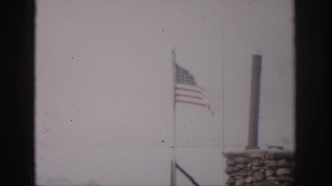 1959 Colorado 8mm frame showing a U.S. flag near a stone chimney, viewed through a dark window frame against a pale sky.