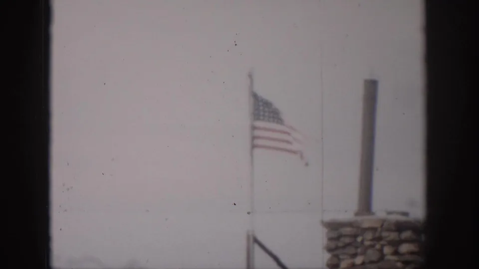 1959 Colorado 8mm frame showing a U.S. flag near a stone chimney, viewed through a dark window frame against a pale sky.