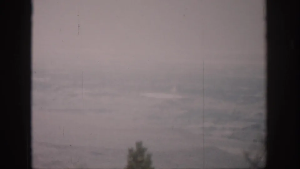 1959 Colorado 8mm frame with distant hazy terrain, pale sky, and small tree tops viewed through a window.