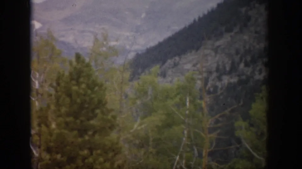 1959 Colorado 8mm view of pine and aspen trees before hazy, rocky mountain slopes, framed by a dark window edge.