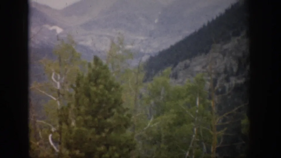 1959 Colorado home-movie frame showing foreground trees and hazy mountain ridges through a dark window opening.