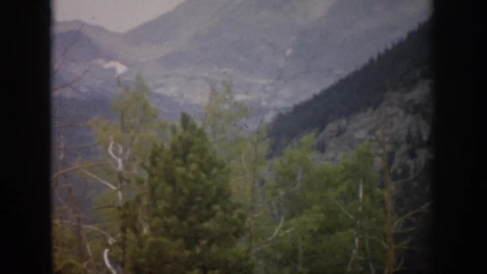 Colorado 8mm frame from 1959 with evergreen and aspen trees in front of hazy mountain terrain seen through a window.