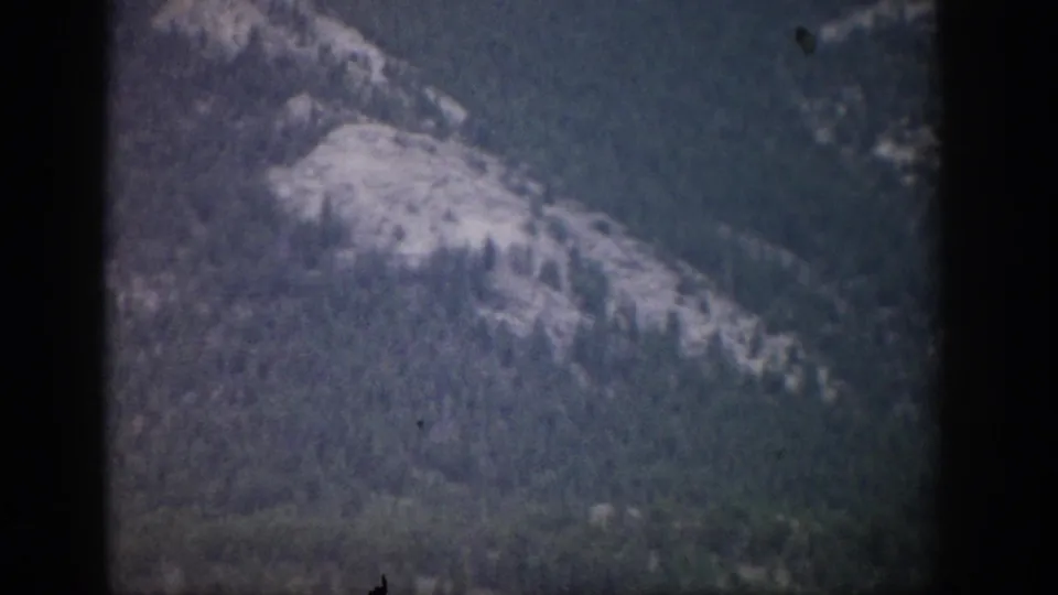 1959 Colorado 8mm frame showing a distant pale rocky slope surrounded by dark forested terrain.