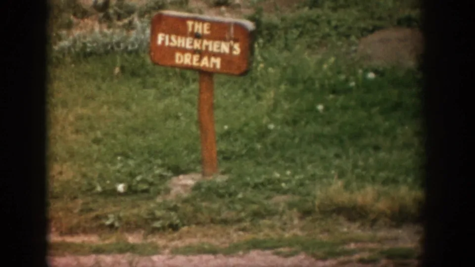 1959 Colorado 8mm frame with the The Fishermen's Dream sign centered near grass, rocks, and a dirt roadside edge.