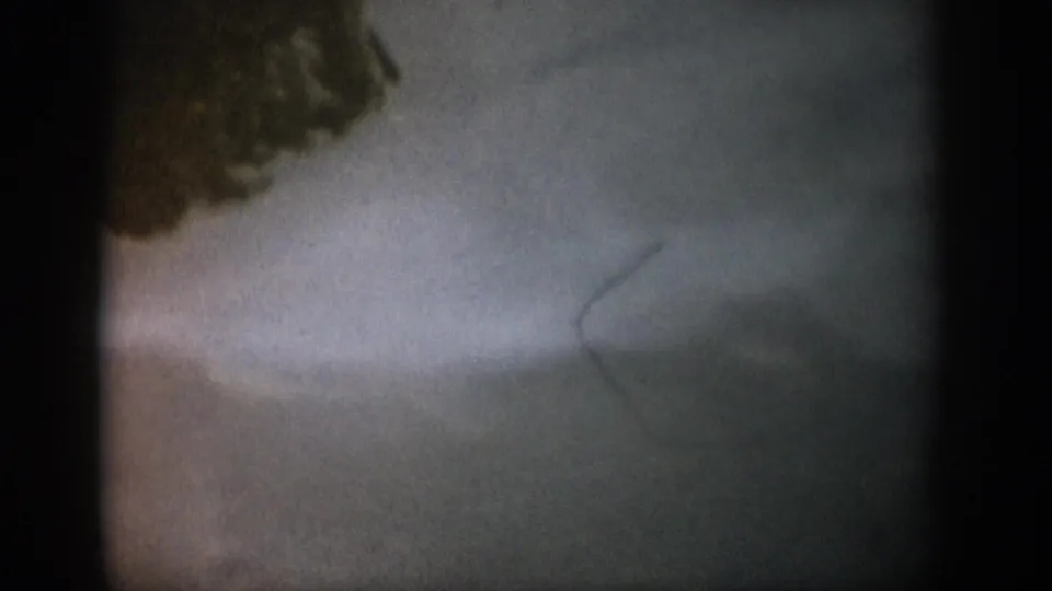 1959 Colorado 8mm frame showing cloudy water near a dark shoreline mass, with a faint fish-like line or tail shape underwater.
