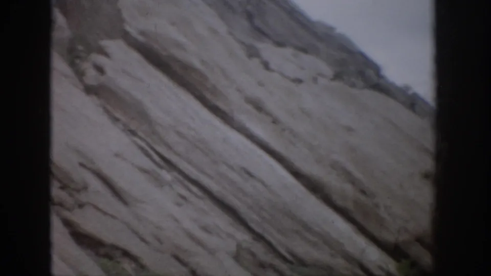 1959 Colorado 8mm frame showing slanted sedimentary rock layers with a small patch of shrubs at the base.