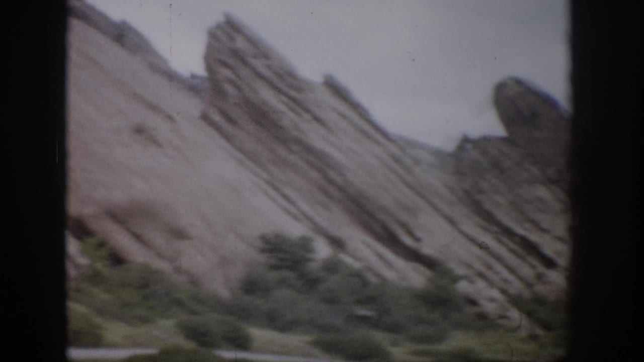 Layered rock formation rises above a road edge and shrubs in a soft 8mm frame.