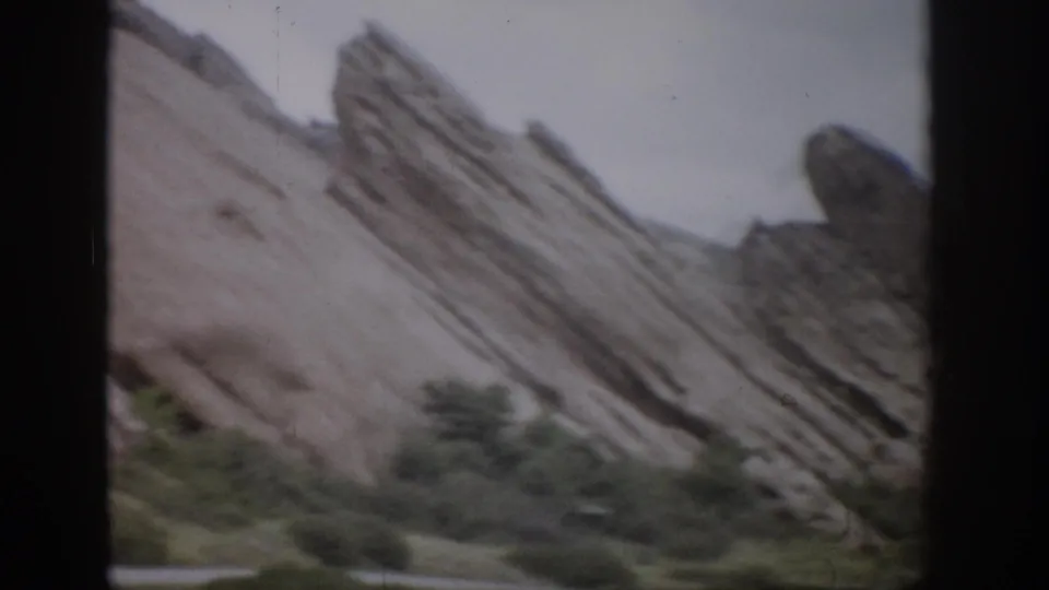 1959 Colorado 8mm frame showing tilted layered rock, roadside greenery, and dark film borders from a visitor viewpoint.