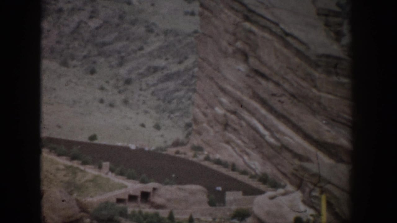 1959 Colorado home-movie frame with a winding road, small roadside structure, and steep layered rock face above it.
