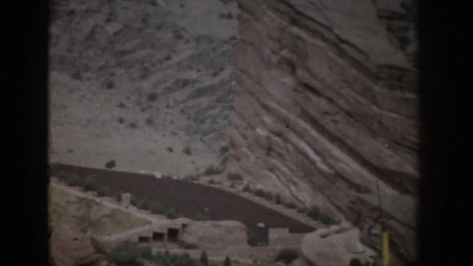 1959 Colorado home-movie frame with a winding road, small roadside structure, and steep layered rock face above it.