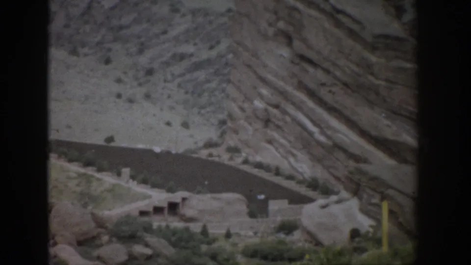 1959 Colorado roadside frame showing layered rock, a curving road below, scattered greenery, and a yellow roadside post near the edge.