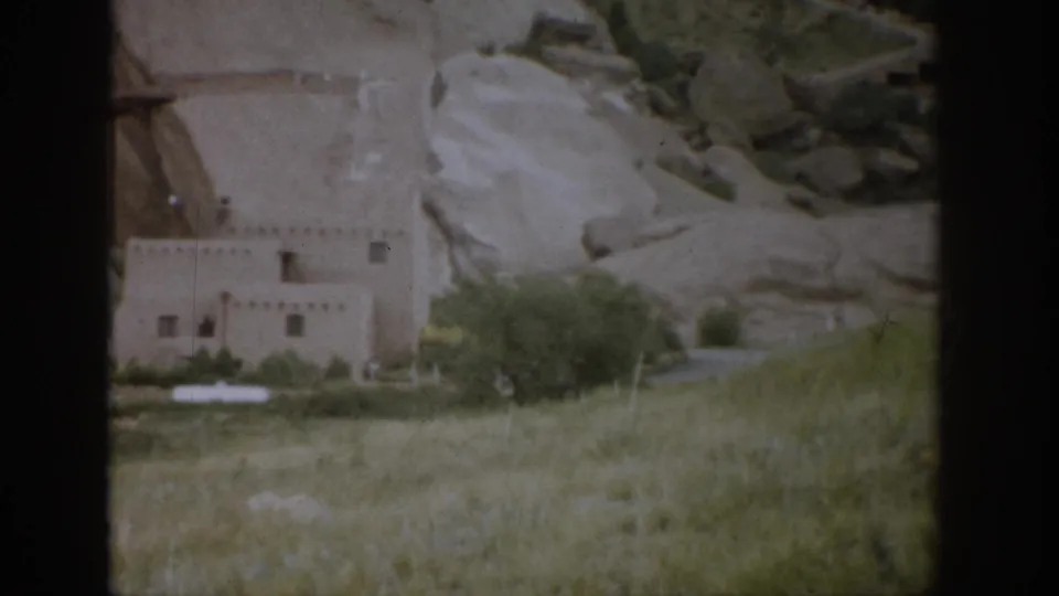 The pan settles on a 1959 Colorado view of an adobe-style building near pale sandstone and scrub vegetation.